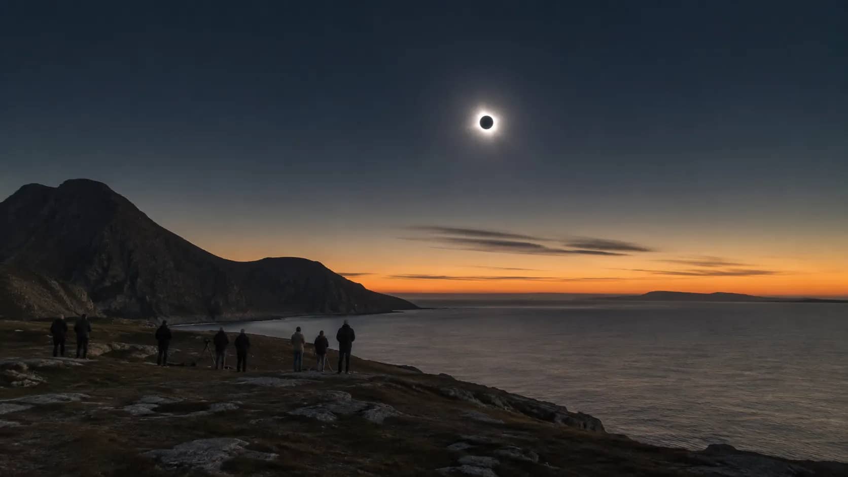 A total solar eclipse above a coastal landscape with the Sun's white corona visible around the dark Moon.