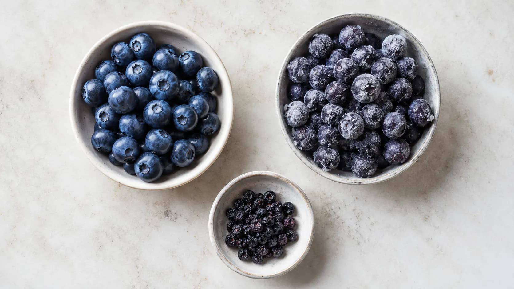 Separate bowls of fresh, frozen and dried blueberries, with a smaller portion of dried fruit.